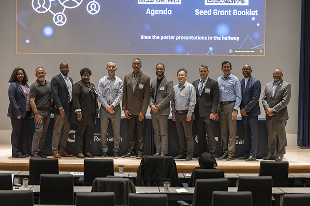 Group of professionals standing on a stage in front of a large presentation screen displaying agenda and seed grant booklet information at a conference.